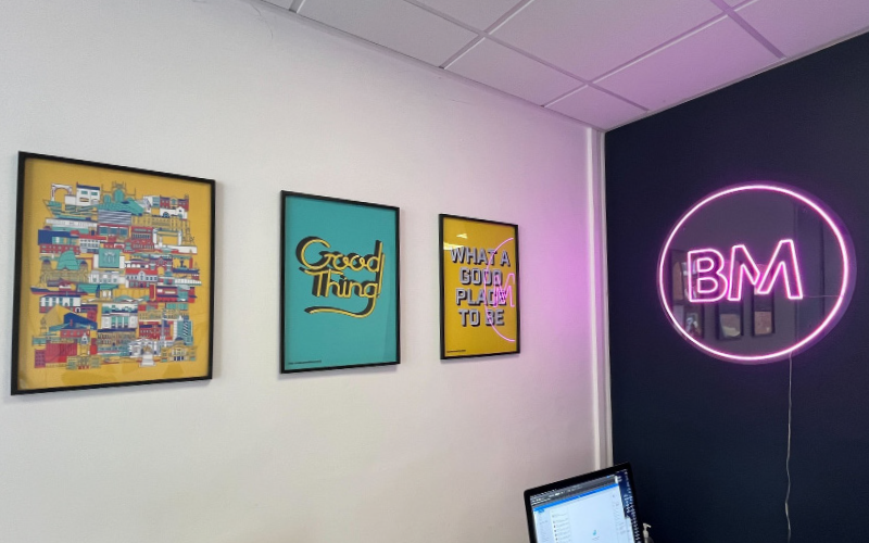 A section of the Bryce Mennell design studio with three framed art prints on the wall, and a neon sign displaying "BM" glowing in pink.