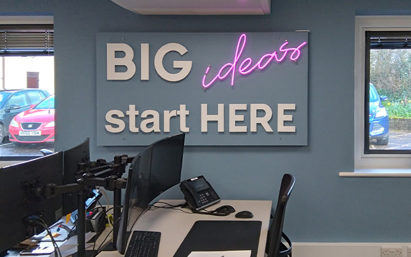 "BIG ideas start HERE" in large, bold letters with "ideas" written in pink neon script. Below is a desk with monitors, a phone, and an office chair.