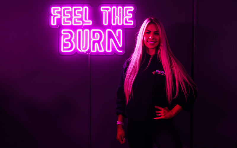 Katie Burns poses in front of a glowing neon sign reading "Feel the burn" in a dark setting.